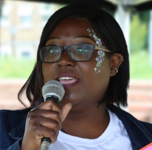 Leila Lawal speaks into a microphone at a community event. She is wearing glasses and has blue floral facepaint on the left side of her face.