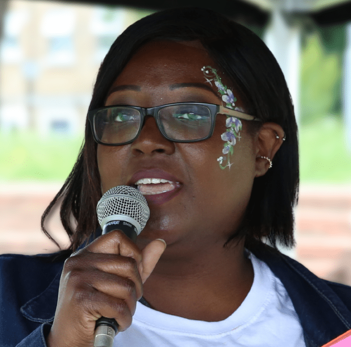 Leila Lawal speaks into a microphone at a community event. She is wearing glasses and has blue floral facepaint on the left side of her face.
