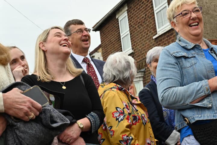 A group of people stand in a group laughing and smiling, with houses behind them
