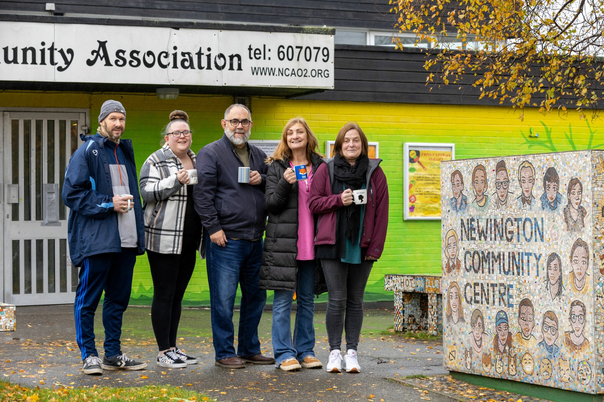 Five people stand in front of a yellow and green painted community building. They hold cups of tea and smile at the camera.