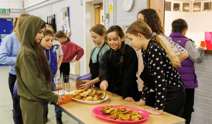 A group of young people at a youth club smile and chat as they stand around a table full of toasted sandwiches they've just made.