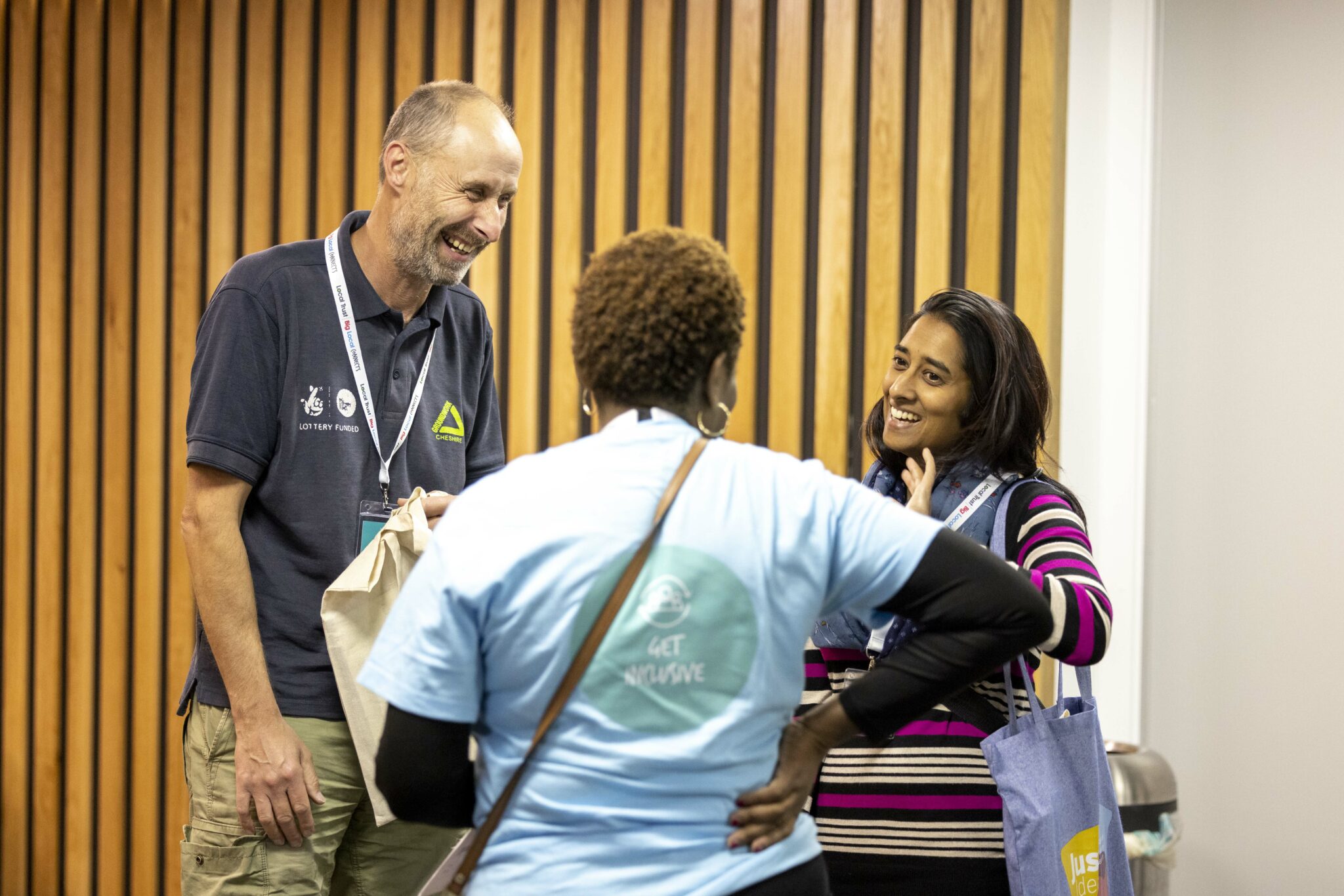 Three people smiling and talking at a conference in an indoor setting. They are wearing white lanyards and there is a wood panelled wall behind them.
