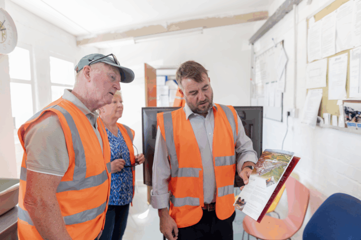 The image shows three people wearing high-visibility orange safety vests inside an office-like setting. One man is holding and pointing to a brochure or document, explaining its contents to the other two individuals, who are attentively looking at it. The room has white walls, bulletin boards covered with papers, and a clock on the wall.