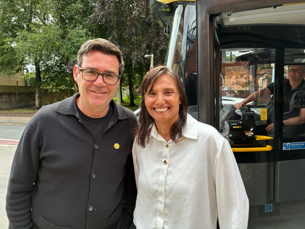 Two smiling individuals standing in front of a Bee Network bus, with a driver visible inside the vehicle. The man on the left wears glasses and a black jacket with a yellow Bee Network badge, and the woman on the right wears a white shirt. Trees and a quiet street are visible in the background.