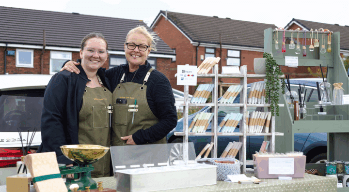 Two smiling women are standing behind a market stall at an outdoor event. They are both wearing matching olive-green aprons labeled 
