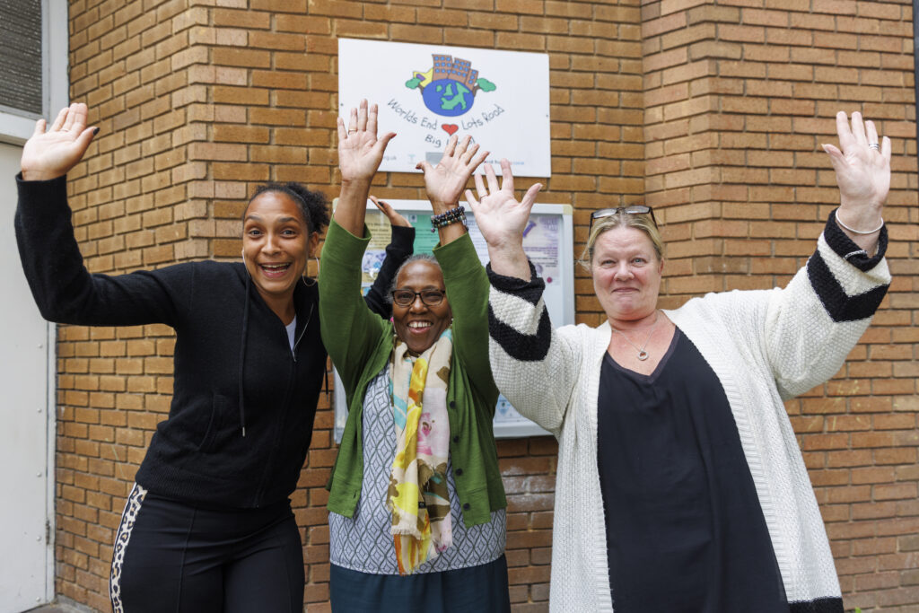 Three women stand outside a brick building, smiling and raising their hands joyfully in the air. Behind them is a white sign with colorful graphics and the text “World’s End Big Local.” The woman on the left wears a black zip-up jacket and patterned leggings. The woman in the middle wears glasses, a green cardigan, and a floral scarf. The woman on the right wears a white cardigan with black stripes and a black dress. The atmosphere is cheerful and welcoming.