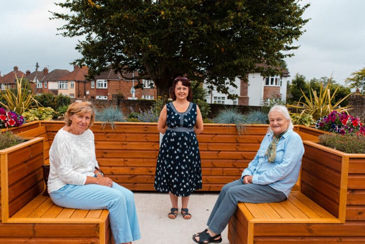2 women sitting and 1 standing to take photo at the community garden