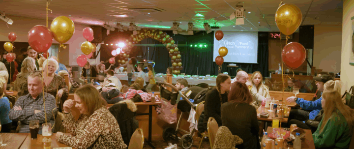 The image shows a lively indoor celebration event with many people seated at tables and children dancing near a stage. The venue is decorated with red and gold balloons, some of which read 