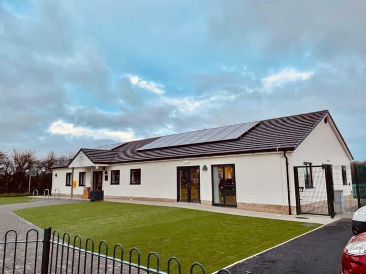 Exterior view of a modern, single-storey community building with a pitched roof and solar panels. The cream-coloured structure features large windows and glass doors, with a well-maintained artificial grass area and a paved path in front. A fenced entrance and car park are visible, with overcast skies and trees in the background.