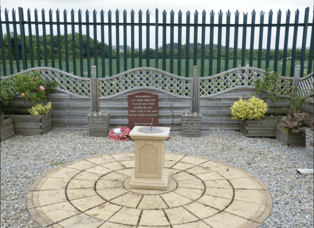 A stone paved circle surrounds a central fountain, creating a serene and inviting outdoor space. There is a brown plaque mounted on the fence behind.