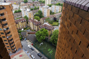 An aerial view of an urban residential area shows many light-brown brick buildings with some red-tiled roofs. In the center, surrounded by buildings, is a green sports court with white lines and a white goal. Cars are parked on the streets around the court. The top of a tall, modern, reddish-brown brick building is visible on the right, and the side of another tall, reddish-brown brick building with many windows and balconies is on the left. In the background, more buildings and trees can be seen.
