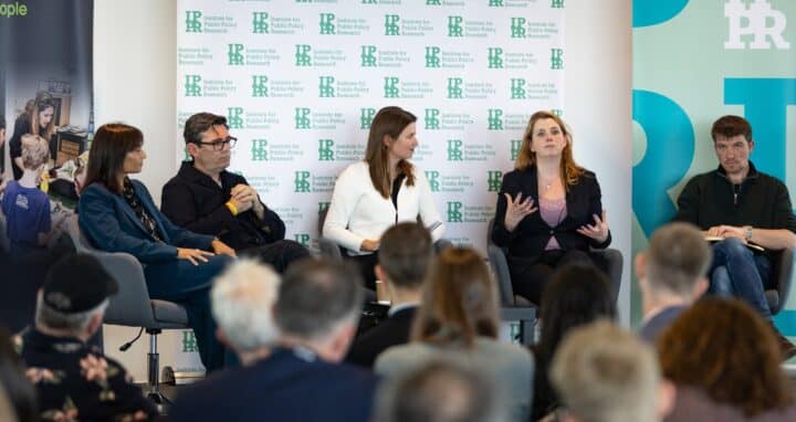 A panel of five people sits on stage in front of a backdrop with the Institute for Public Policy Research (IPPR) logo. They are engaged in discussion, with one woman speaking while the others listen attentively. The audience is visible in the foreground, watching the conversation. Bottles of water and notebooks are on the table between the panelists.