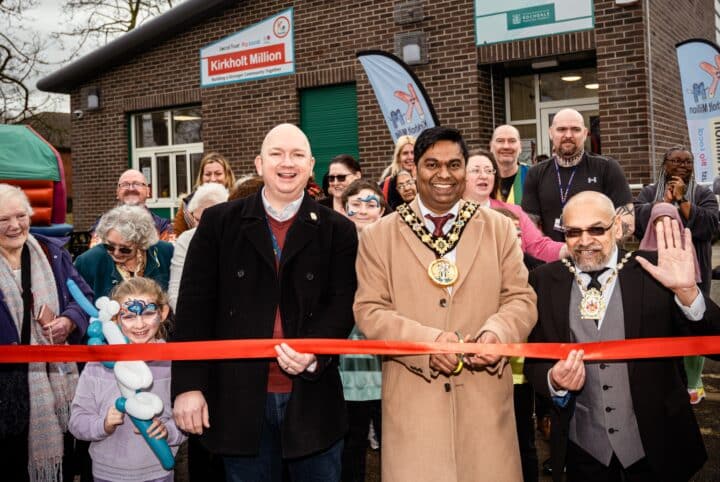 A group of people gathered outside a community building during a ribbon-cutting ceremony. Three officials, one holding scissors, stand at the front smiling, surrounded by attendees including children and elderly community members.