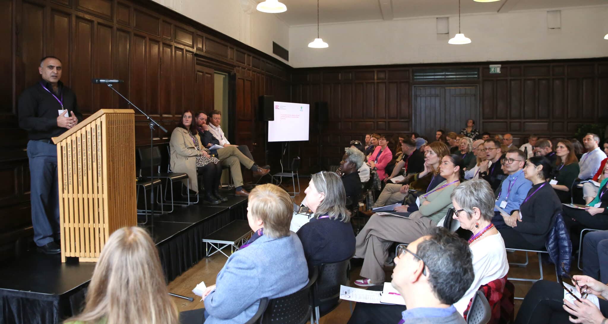 A man stands at a podium speaking to an audience of conference attendees. They are sat in rows in a wood panelled room. Three other speakers are sat on the stage to the right of the podium,