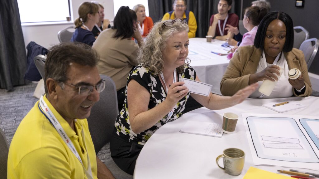 Attendees engage in a group activity at a round table covered with worksheets, pens, cups and cards. On the left, a man in a yellow polo shirt with glasses smiles. In the centre, a woman with curly grey-blonde hair in a black floral top holds a card and gestures as she speaks. On the right, a woman in a tan blazer examines a card. Other attendees are visible at tables in the background