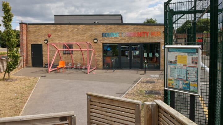 A single storey brick building with three panelled glass doors and windows. A sign above the doors reads 'Bourne Community Hub'. There is a red bike rack to the left of the doors.