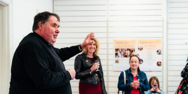 A man in black clothes gestures while speaking to a small group in a bright indoor space, with three adults and a child listening behind him, some holding drinks. Two posters on the wall behind them read “From Post to Purpose” at the top, with smaller text and photos beneath.